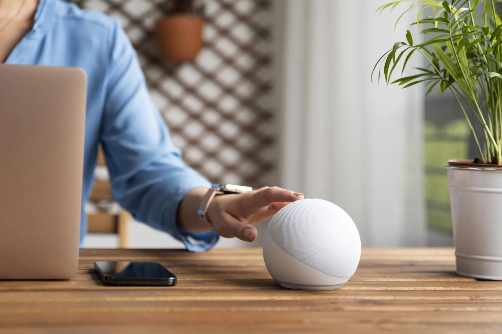 Eine Hand tippt auf einen kugelförmigen weißen Smart Speaker, der auf einem Holztisch neben Laptop und Smartphone steht.